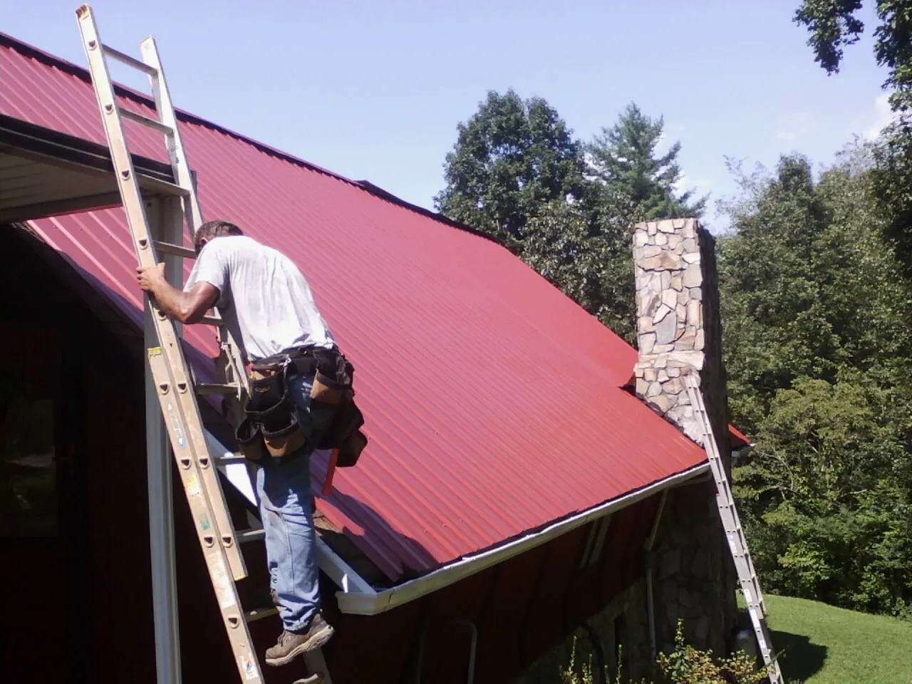 Roofer on a ladder beside a red metal roof and stone chimney — Soffit & Fascia Repair by Roofing Brothers in Millbury